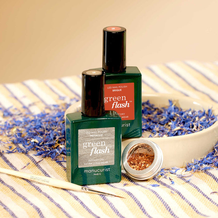 Two bottles of Manucurist Oriental Vibe nail polish and a pot of gold biodegradable glitter from the Autumn 2023 collection sit in a beige bowl on a striped cloth, surrounded by dried blue and purple flower petals. A manicure brush lies nearby.