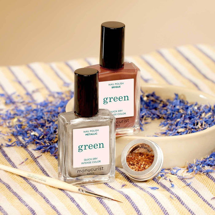 Two bottles of Manucurist Oriental Vibe nail polish (metallic silver and brick red) from the Autumn 2023 collection and a small open jar of biodegradable glitter rest on a plate with dried blue petals, on striped cloth. A brush lies beside them.