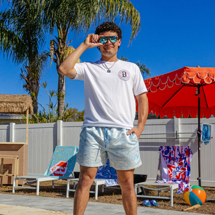 Male model wearing BL101 blue paisley swim shorts and white tee, posing poolside with sunglasses.