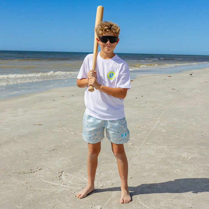 Young boy wearing BL101 blue paisley swim shorts and white tee, holding a baseball bat on the beach.