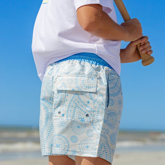 Rear view of BL101 youth blue paisley swim shorts on a boy holding a baseball bat at the beach, showing the back cargo pocket with drain grommets.