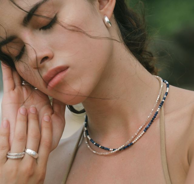 Woman with a calm expression, eyes closed, showcasing silver rings and stacked beaded necklaces from luna rose