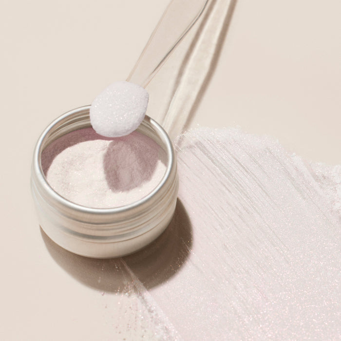 A small metal jar of Manucurist Pink Glazed Effect Powder, filled with fine, white powder with a subtle pearlescent sheen, sits on a light surface. A clear plastic spoon holds some of the shimmering powder beside the jar.