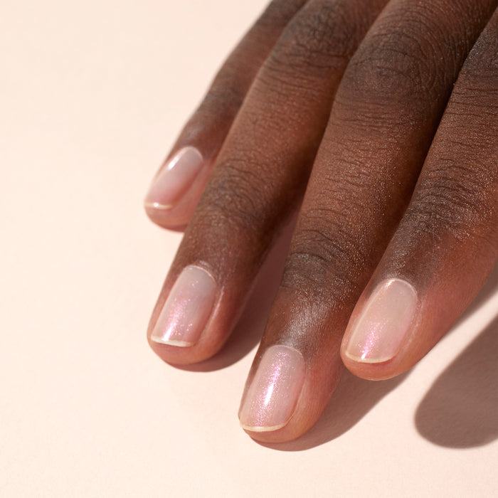 A medium-dark skinned hand with neatly trimmed nails shows off light pink, pearlescent Manucurist Pink Glazed Effect Powder polish, resting on a pale neutral background.