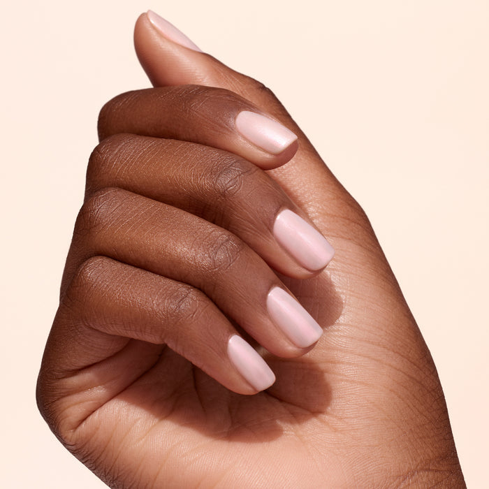 A hand with medium brown skin tone is shown against a light beige background. The nails are neatly manicured, painted pale pink, and topped with Manucurist Pink Glazed Effect Powder for a subtle, pearlescent sheen.