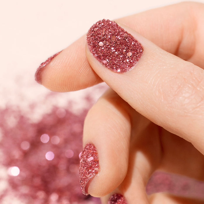 A close-up of a hand with nails featuring Manucurist's Pink Quartz Biodegradable Glitter, stunning nail art, and sparkly accents scattered in the background.