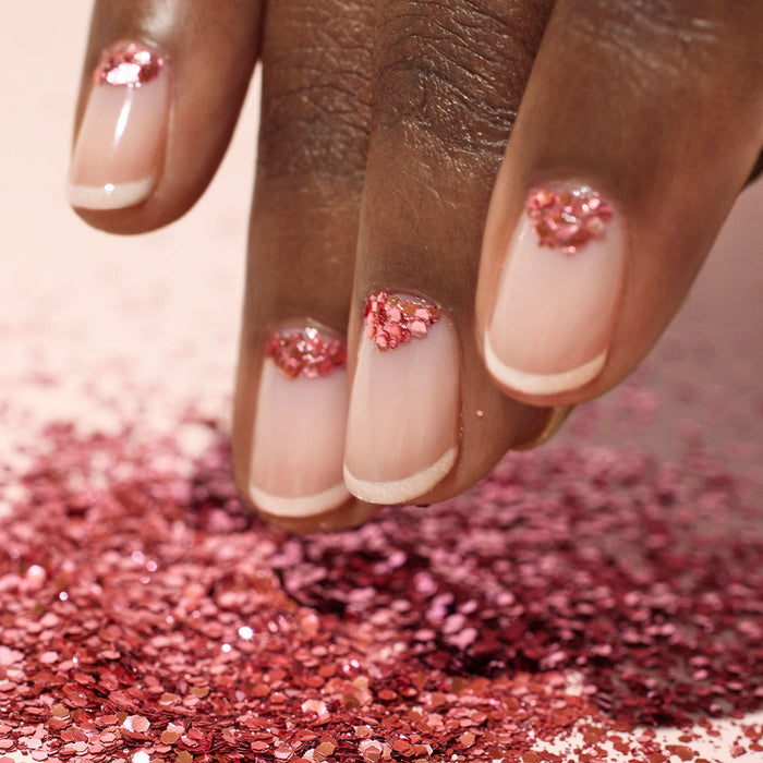 A close-up of a hand with nude French manicure nails, each adorned near the cuticles with Manucurist Pink Quartz Biodegradable Glitter. The pink glitter is also scattered below, accentuating the elegant nail art.