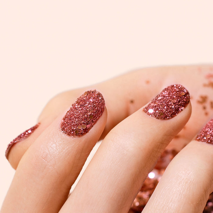 A close-up of a hand with nails painted in sparkly pink, featuring stunning nail art and shimmering Manucurist Pink Quartz Biodegradable Glitter scattered in the background.