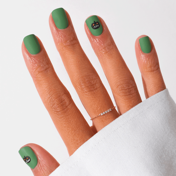 A hand with Pumpkin Patch matte green gel polish by Manucurist, featuring small black cat faces on the pinky and thumb. The look is finished with a thin gold ring and a white long-sleeve shirt—perfect subtle Halloween nail art.