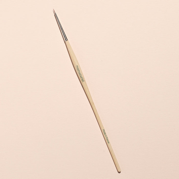 The Manucurist Pumpkin Patch paintbrush, featuring a thin wooden handle and fine pointed tip ideal for detailed Halloween nail art, is placed diagonally on a light beige background.