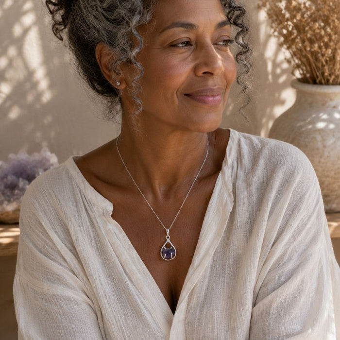 Woman wearing a raw amethyst teardrop pendant in a polished silver setting on a delicate chain with a relaxed linen blouse in soft natural light.
