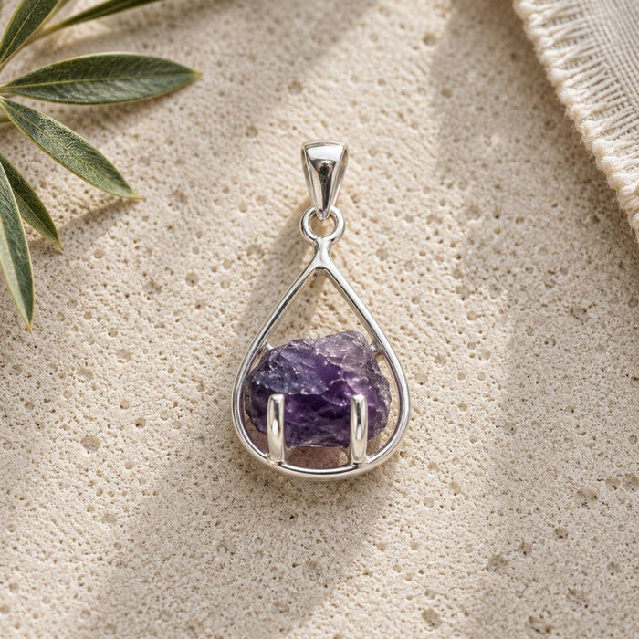 Raw amethyst teardrop pendant in a polished silver setting displayed on a natural stone surface with soft daylight and botanical accents.