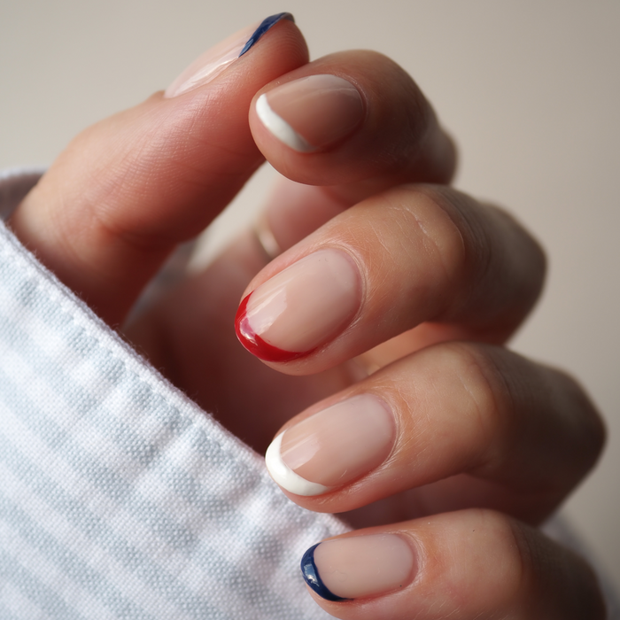 A hand with a neutral manicure using the Manucurist Red, White  Blue Trio—blue on pinky and thumb, red on middle, white on index and ring—paired with a striped sleeve and matching bold red, white, and blue eyeshadow.