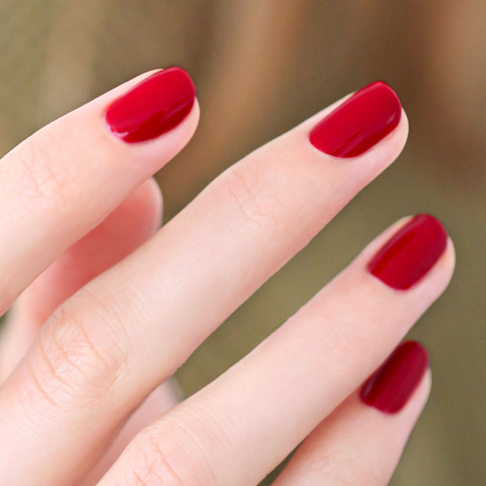 A close-up showcases neatly manicured nails coated in glossy dark red, featuring Manucurist’s Red, White  Blue Trio. The blurred background subtly highlights the trio's red, white, and blue shades for a patriotic pop of color.