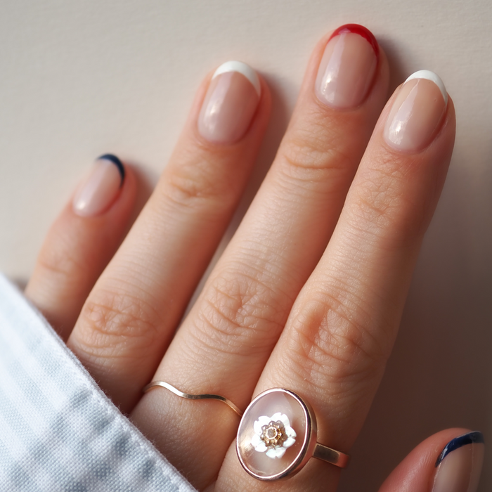 A hand with nails painted in Manucurist's Red, White  Blue Trio—red, navy blue, and white—echoes the bold flair of patriotic eyeshadow. Two gold rings adorn the fingers: one slim, the other featuring a round floral pattern.