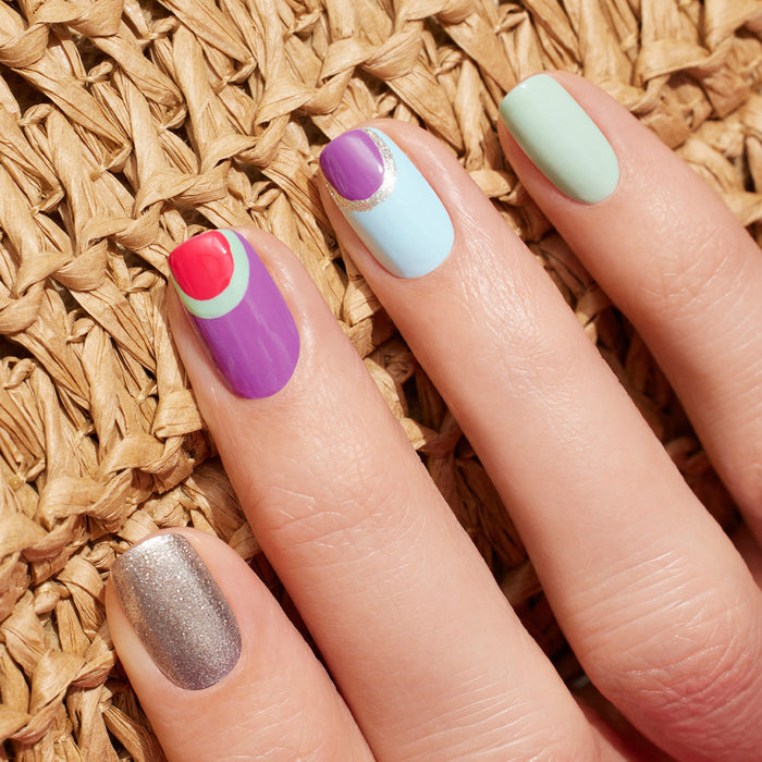 A hand showcases Manucurist's Retro Rainbow nails: silver glitter, purple with red and green shapes, blue with a purple tip and silver stripe, plus solid Green Flash pastel green—all from the Summer Love collection—resting on a woven surface.