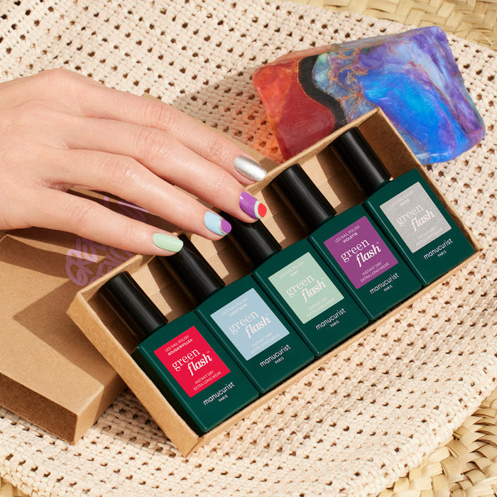 A hand with Manucurist's Retro Rainbow nail art holds a box of five Green Flash polishes, neatly arranged on a woven surface alongside a multicolored stone and a sun hat.
