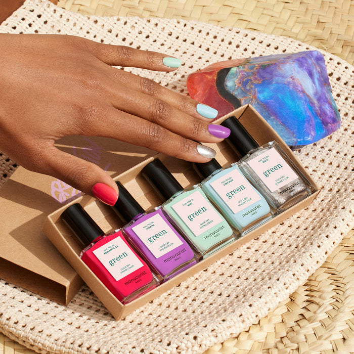 A hand with multicolored nails rests on a woven mat beside an iridescent stone, holding Manucurist's Retro Rainbow box—five pastel polishes ideal for creating vibrant nail art.