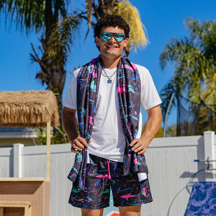 Male model wearing BL101 neon shark swim shorts and white tee poolside, with a matching shark print towel draped over his shoulders.