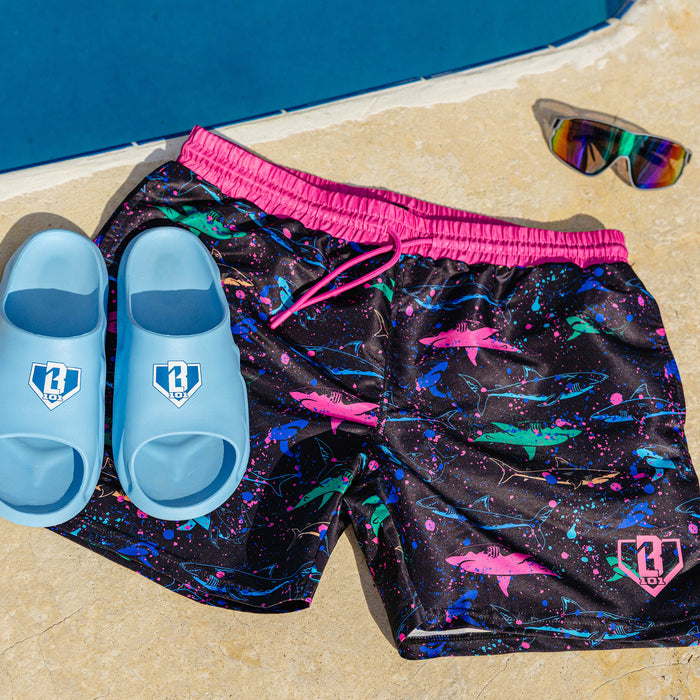BL101 black neon shark swim shorts laying on a ground next to glasses and blue BL101 slides