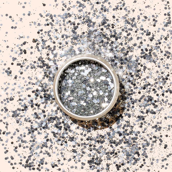 A round container of Manucurist Silver Biodegradable Glitter rests on a beige surface, surrounded by scattered silver sparkles—ideal for eco-friendly nail polish or festive silver nail art.