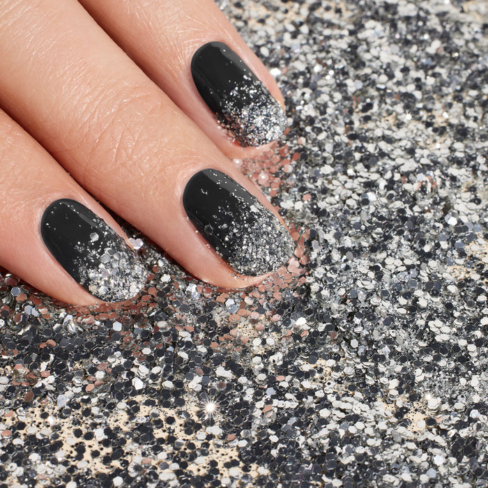 A hand with black nails and silver ombre tips, created using Manucurist Silver Biodegradable Glitter and eco-friendly polish, rests on a surface sprinkled with sparkling silver glitter.