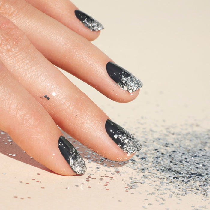 A hand with black nails featuring silver gradient nail art at the tips rests on a surface sprinkled with Manucurist Silver Biodegradable Glitter.