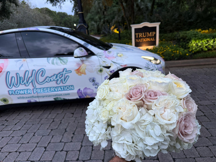 Bouquet of flowers held in front of a vehicle with 'Wild Coast Flower Preservation' branding, and a 'Trump National' sign in the background.