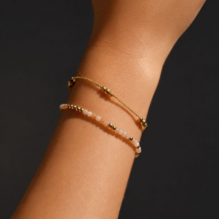 Two gold beaded bracelets on a wrist against a dark background