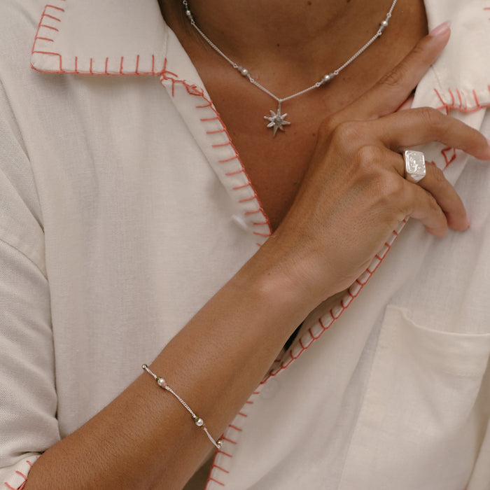 Close-up of a person wearing a silver necklace, bracelet, and ring on a light-colored garment with red stitching.