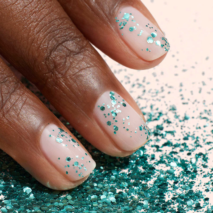 A close-up of a hand with natural nails adorned with Manucurist Turquoise Biodegradable Glitter, surrounded by matching loose glitter on a light surface.