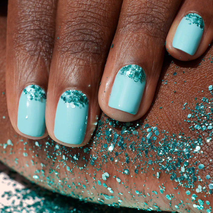 A hand with aqua blue polish and turquoise nail art rests on skin dusted with Manucurist Turquoise Biodegradable Glitter.